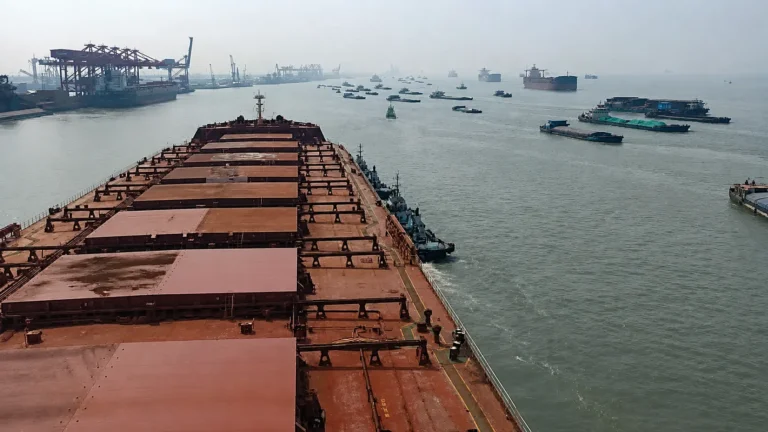 Bulk carrier approaching a busy port area with vessels waiting and terminal cranes in the distance