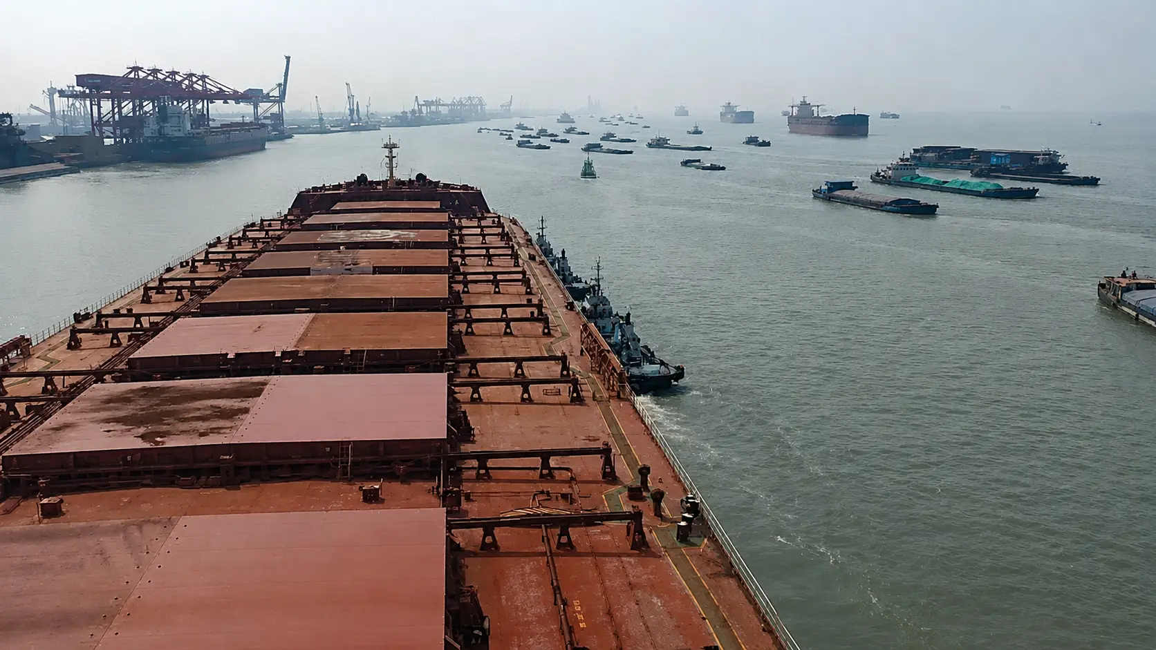 Bulk carrier approaching a busy port area with vessels waiting and terminal cranes in the distance